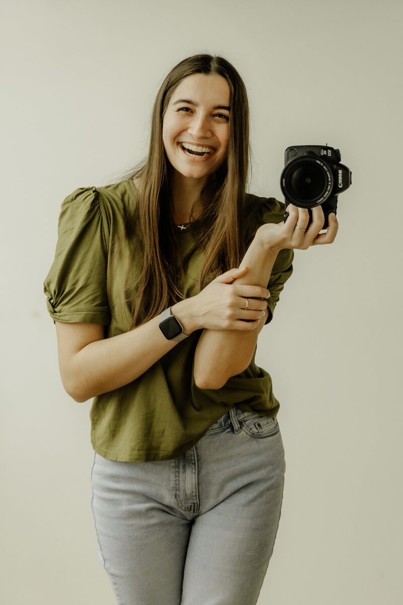 Professional wedding photographer smiles at the camera as she stands in an open field while wearing a lovely gingham dress.