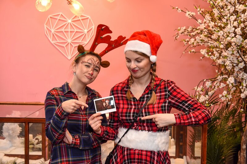 Two women in plaid outfits posing in a holiday-themed photo booth, wearing reindeer and Santa hats while holding a printed photo as a keepsake.