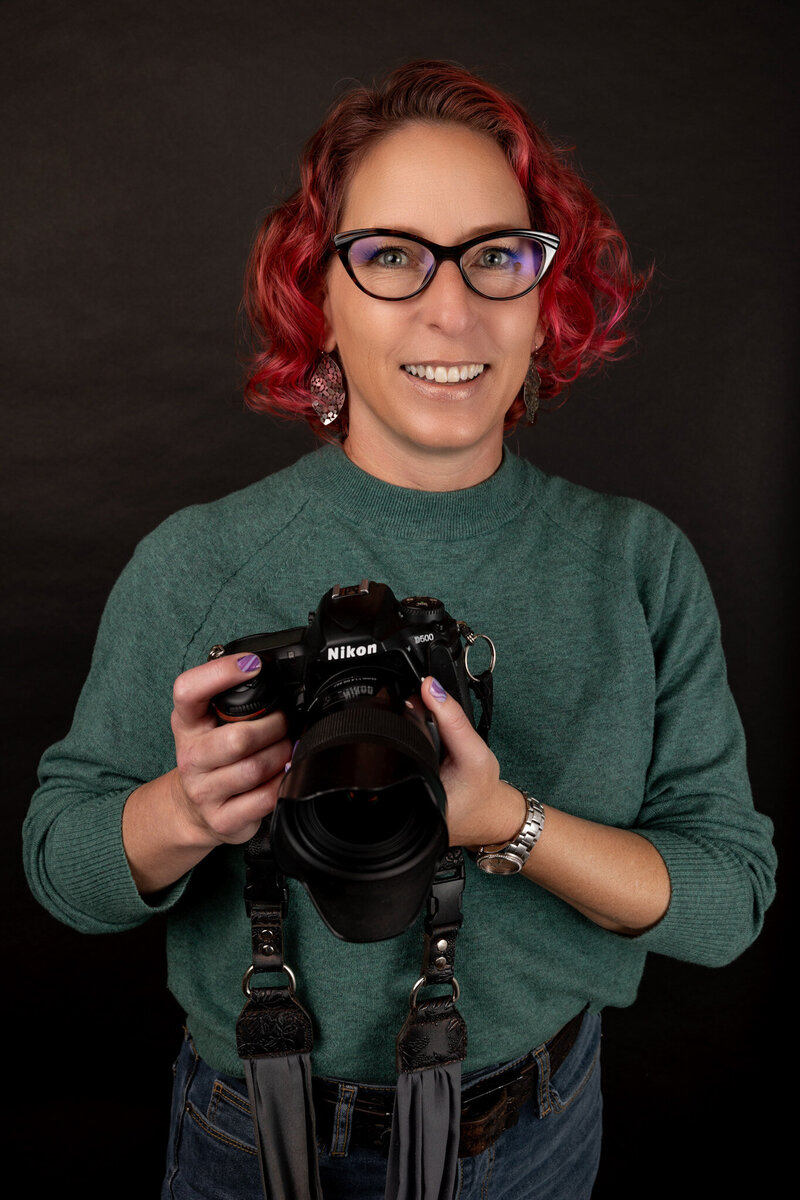 headshot of woman with glasses and pink hair wearting green sweater holding camera