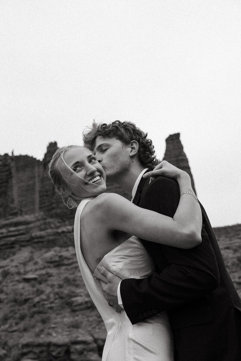Husband kissing wife on cheek in black and white while wearing their wedding attire with rocks in the background 