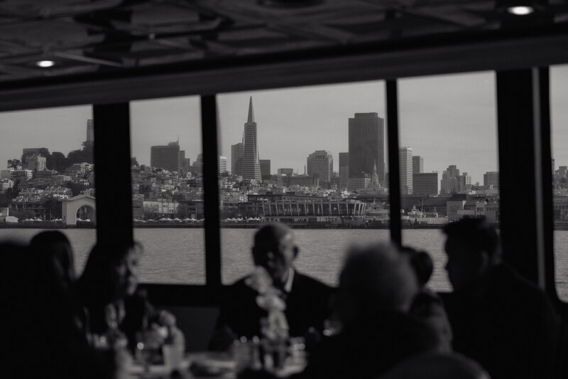 Guests dining onboard yacht with iconic San Francisco cityscape visible through windows, intimate documentary photography.