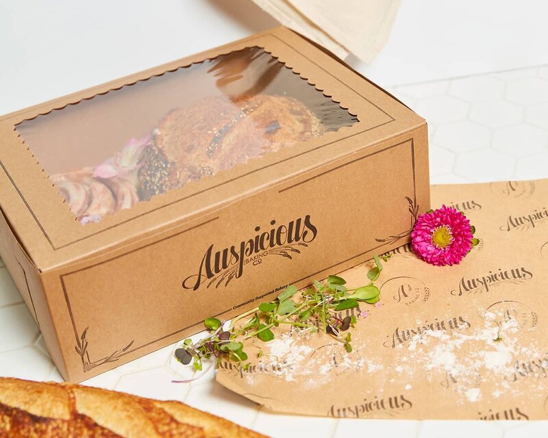 A brown, kraft bakery box filled with pastries visible through the window on the fop of the box. The front is printed with the Auspicious logo and a custom design