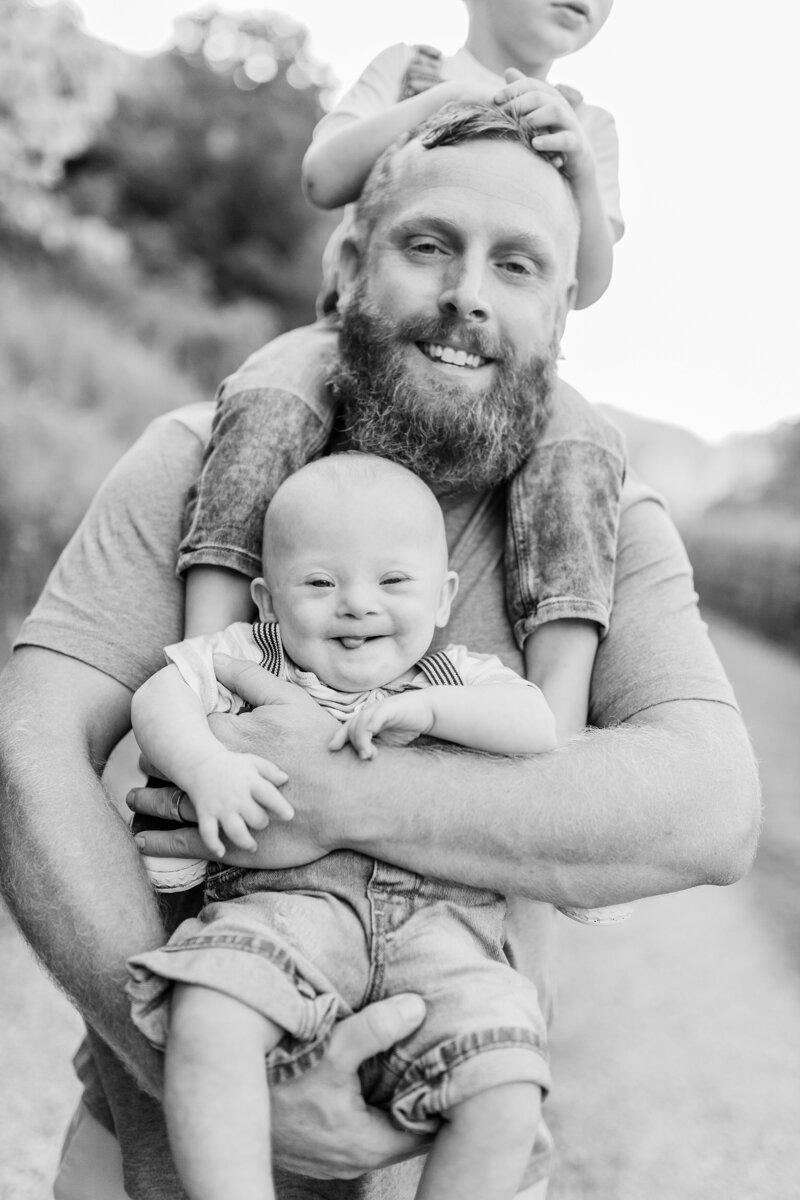 A bearded Dad holding a son on his shoulders and another in his arms while they laugh at photo session in Lexington KY.