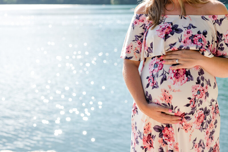 Pregnant woman in floral dress holding stomach in front of a lake