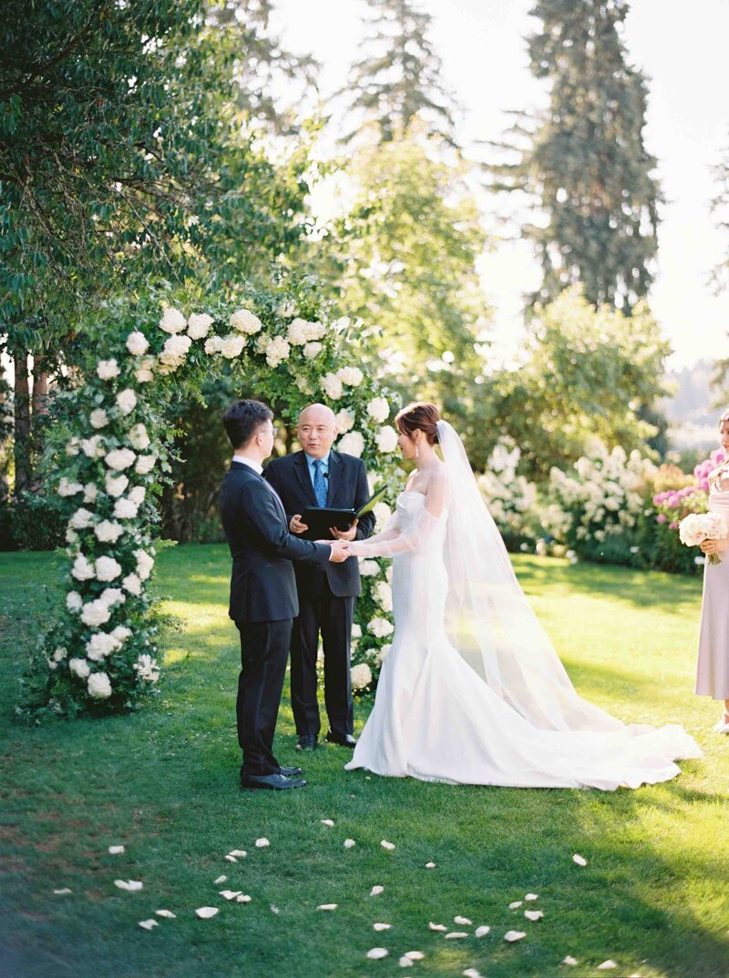 Seattle Wedding Photographers lush green garden wedding ceremony with white rose arch and couple holding hands