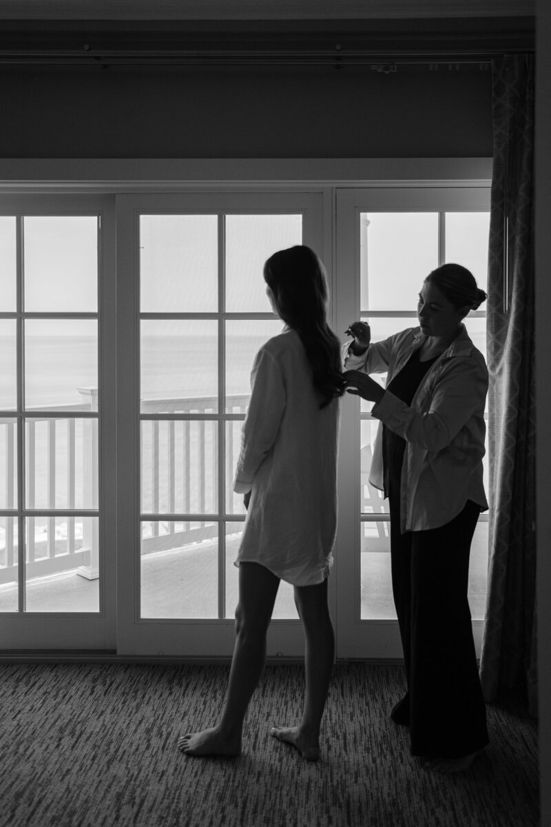 Bride in a robe being styled by her hairstylist in front of large balcony doors