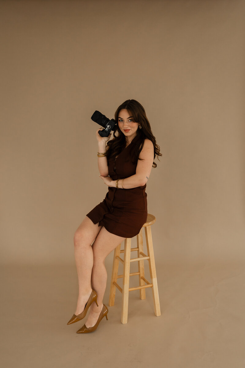 Abby Dutra sitting on a stool in her photography studio, smiling warmly in a relaxed and professional setting.