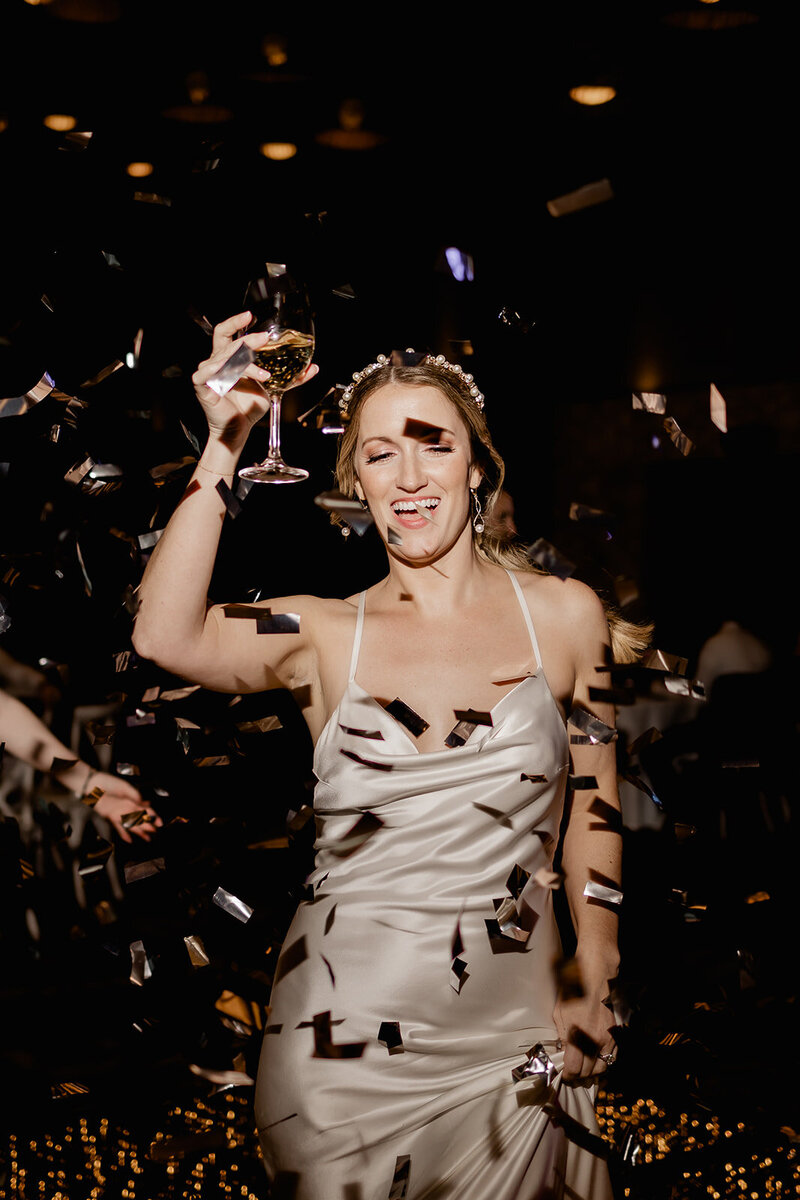 A bride celebrates on the dance floor, holding a glass of wine and smiling as confetti falls around her. She is wearing a silk gown and a jeweled headpiece, captured in a fun, lively moment