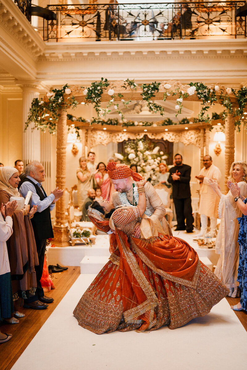 A stunning moment from an Indian wedding at the grand Main Hall of Hedsor House in Buckinghamshire. The Polish groom, dressed in traditional Indian wedding attire, gracefully dips his Indian bride, who shines in a breathtaking rustic and gold wedding lehenga. She smiles in his arms as wedding guests cheer and clap in delight. Behind them, the beautifully decorated mandap stands as a symbol of their union, adding to the magic of this joyful celebration.
