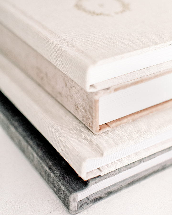 Linen heirloom photo albums stacked on a white table, highlighting custom artwork from Charlotte newborn photographer.