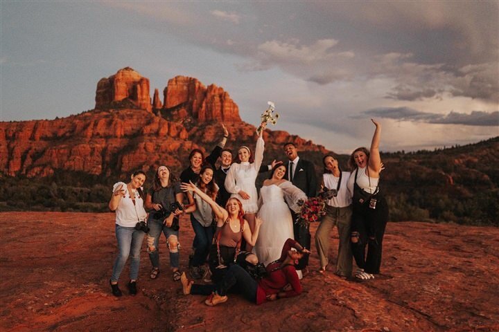 A joyful group poses with a couple at a scenic desert location in Sedona, Arizona, with striking red rock formations in the background. The group enthusiastically celebrates, with the couple at the center holding a bouquet.