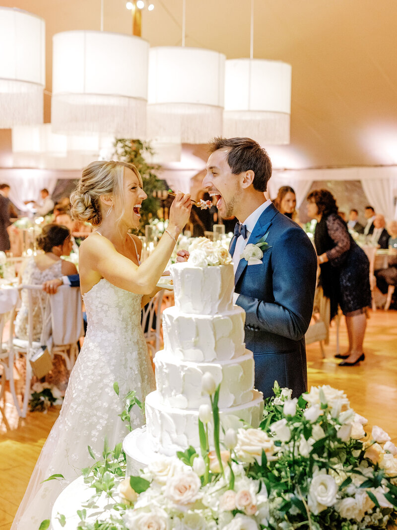 Bride and groom smiling and feeding each other cake in front of their large tiered wedding cake