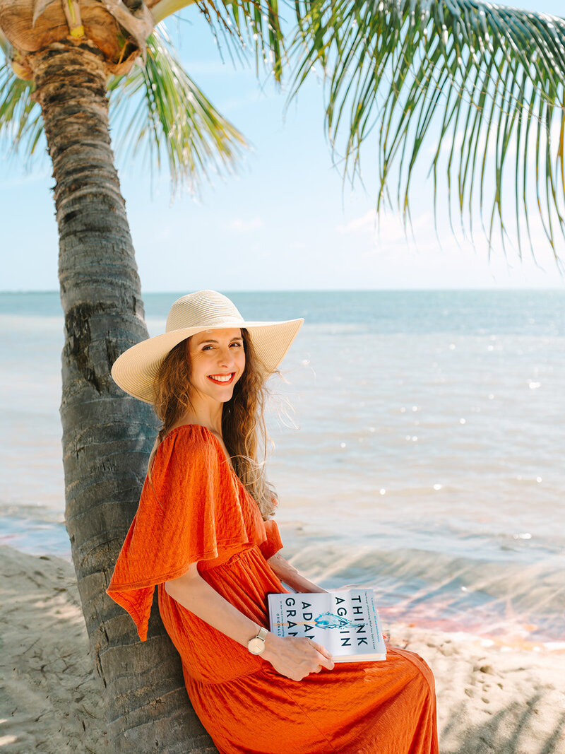 Kilee in an orange dress and straw hat sitting on a palm tree with a book in front of the ocean