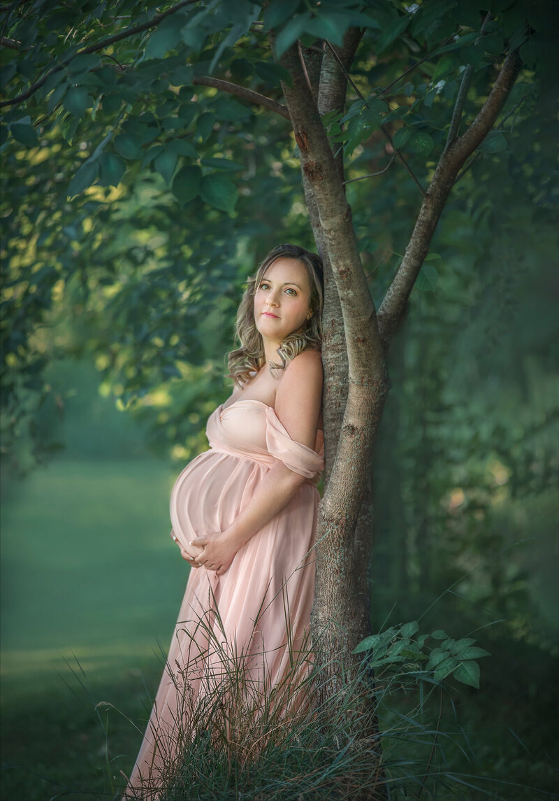 "Pregnant mom-to-be in a flowing pink gown, gently leaning against a tree, captured in a serene and natural maternity photography session in Ottawa."