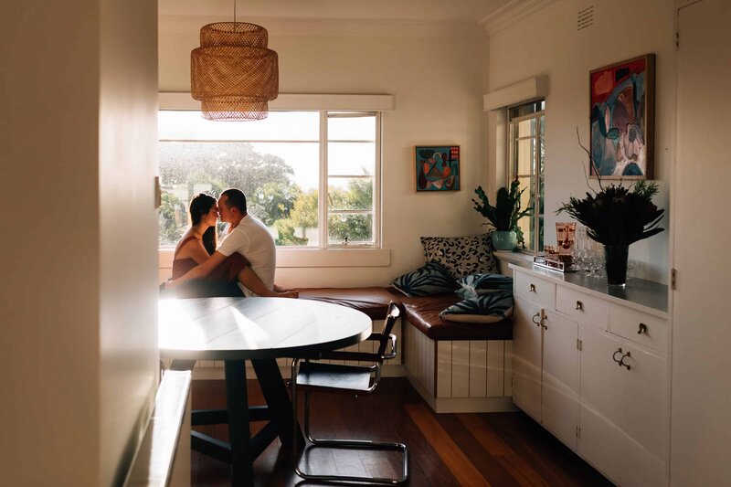 Couple sitting on bench, entwinned, in front of window with light streaming in, during couple photoshoot by And So I Don't Forget Photography Melbourne