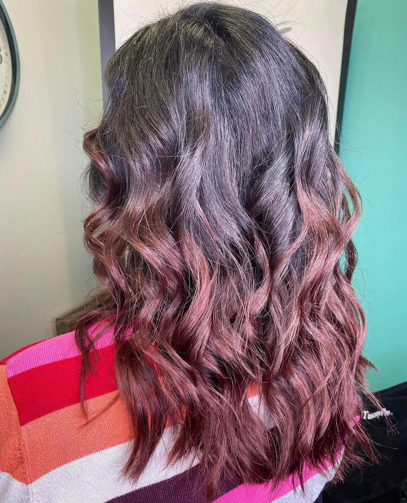 A back view of a woman with dark hair transitioning to vibrant waves of color, showcasing an ombre hairstyle created by 212 Salon, Spa, & Barbershop.