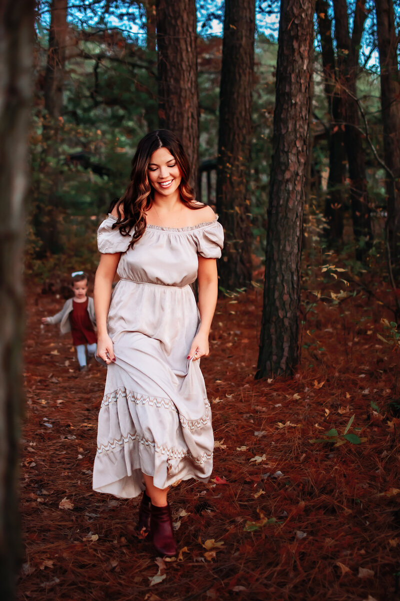 A woman is walking toward the camera while looking down and smiling in a long flowy tan dress. There's a toddler in the background running behind her. They're in a forested area with trees.