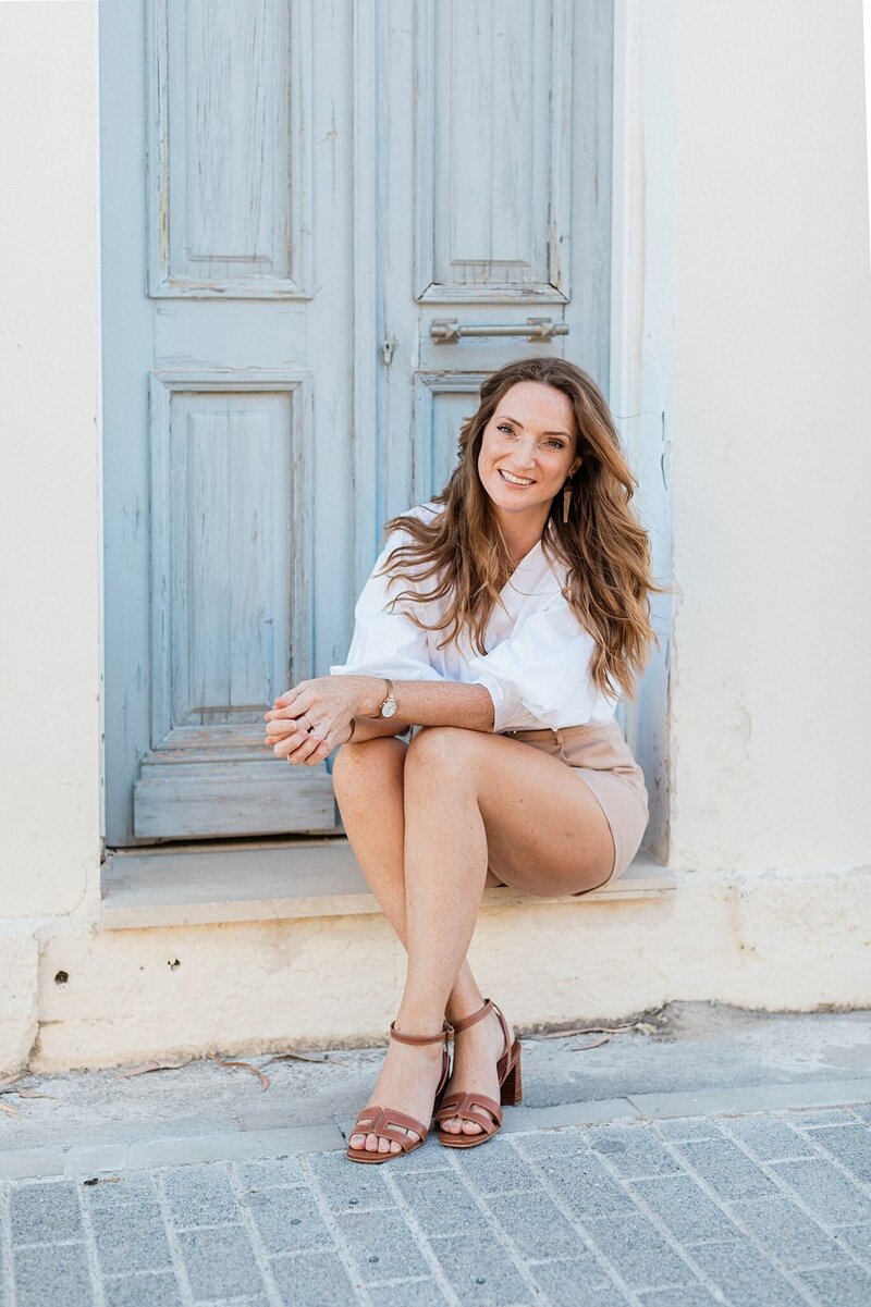 Claire, founder of Lefkas Weddings, smiling in a relaxed and stylish portrait while seated in front of a rustic blue door
