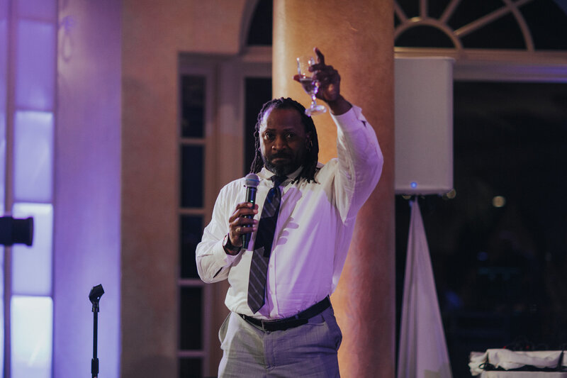 Wedding DJ holding a microphone and raising a glass during a heartfelt toast, with clear, professional audio setup in the background.