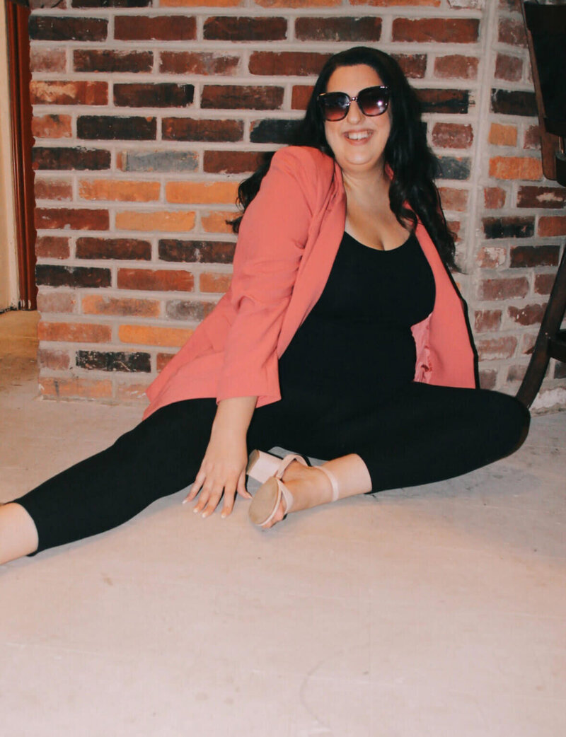 Sarah Weiss sitting against a brick wall, wearing a pink blazer and sunglasses and smiling