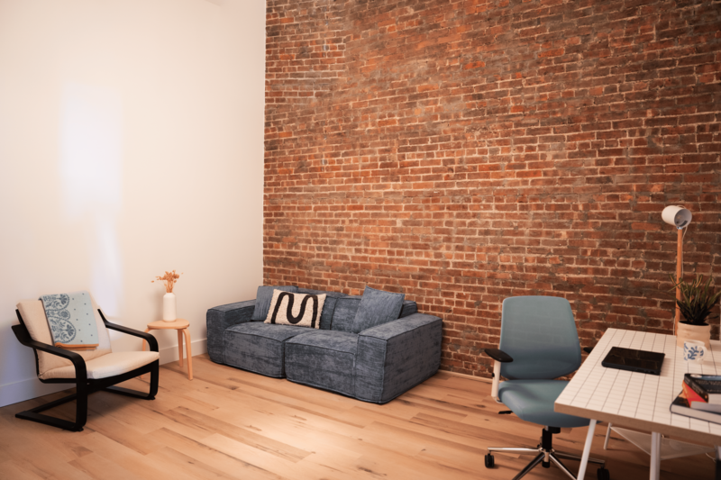 Cozy therapy office with an exposed brick wall and a large, plush blue sofa, chairs and a desk.