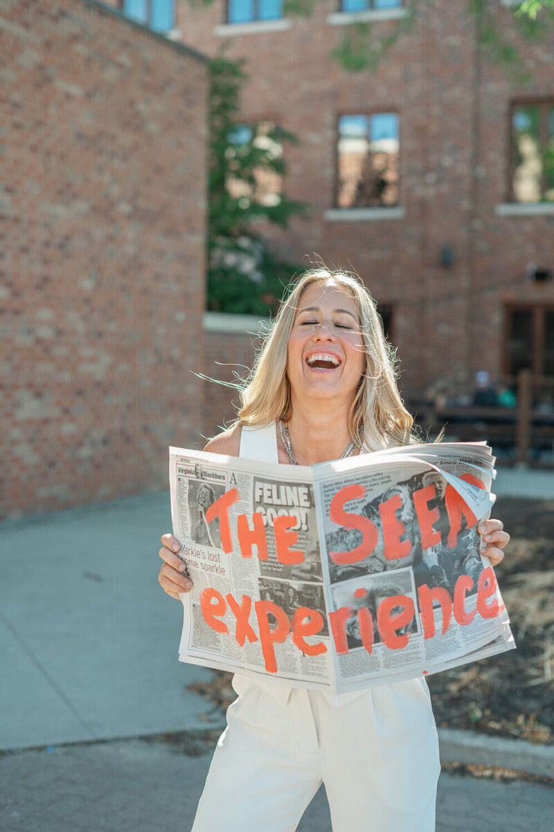 Kristen, Kelowna photographer holding newspaper painted with the words 'the SEEN experience" and laughing.