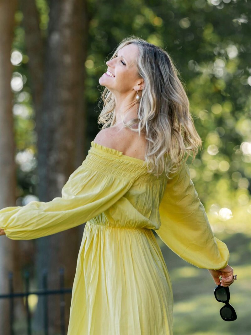 Phillipa Leseberg with long hair in a yellow off-shoulder dress, author of His Name is Eric, smiles while holding sunglasses outdoors in a park-like setting.
