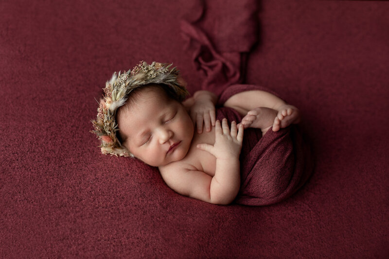 Newborn baby girl posed on a maroon blanket curled up in womb like position with a floral crown
