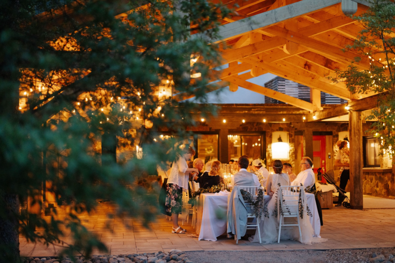 An elegantly set table under a wooden pavilion, decorated with white linens, greenery, and tall glass candle holders. String lights are draped across the wooden beams, and sheer white curtains hang in the background, creating a romantic atmosphere at night seen through trees.