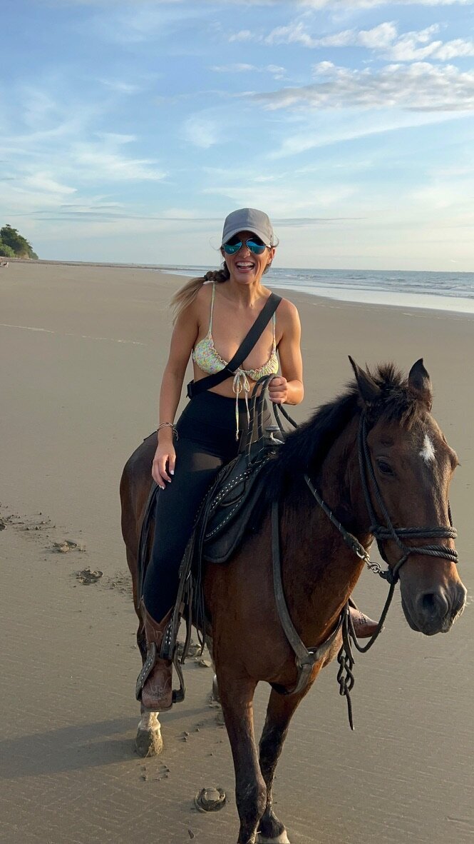 Vanessa Ramirez, owner of Sabri & Co. sits atop a horse on a sunny beach day. She wears sunglasses and a hat while smiling gleefully at the camera.