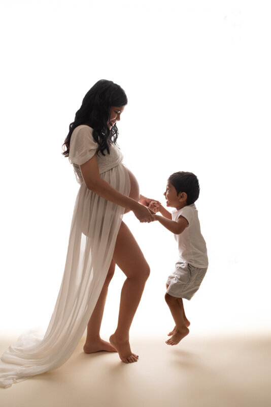 Atlanta, Georgia maternity photography studio session with mom in sheer maternity gown holding 3 year old son's hands looking at each other.
