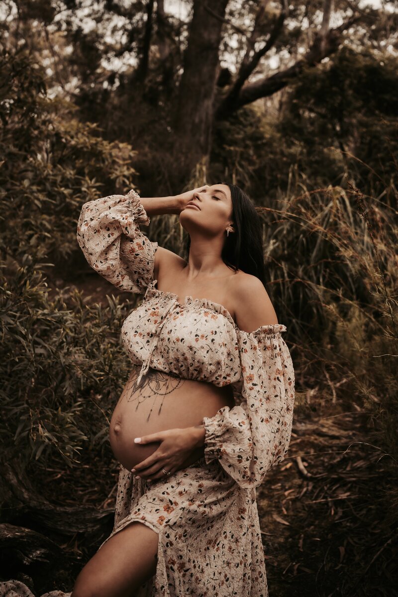 Pregnant woman in floral off-shoulder dress stands in a forest, one hand on her head, the other on her exposed belly—an exquisite moment captured by a Wollongong maternity photographer.