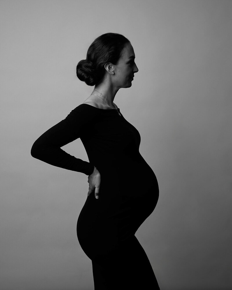 Black and white profile portrait of a pregnant woman standing tall in a fitted black gown with an off-the-shoulder neckline. Her hair is elegantly styled in a low bun, and she rests one hand on her hip while the other gently supports her belly. The soft lighting and clean background highlight her strong silhouette, capturing a moment of grace, power, and maternal beauty.