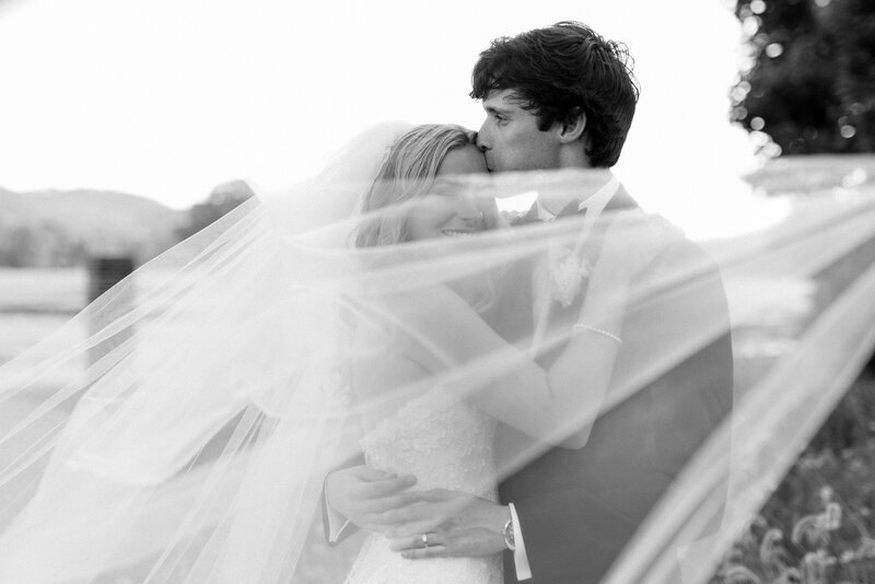 A black and white image of a groom tenderly kissing his bride's forehead as her veil blows across their faces at the Hamilton Pursell Farms in Alabama.