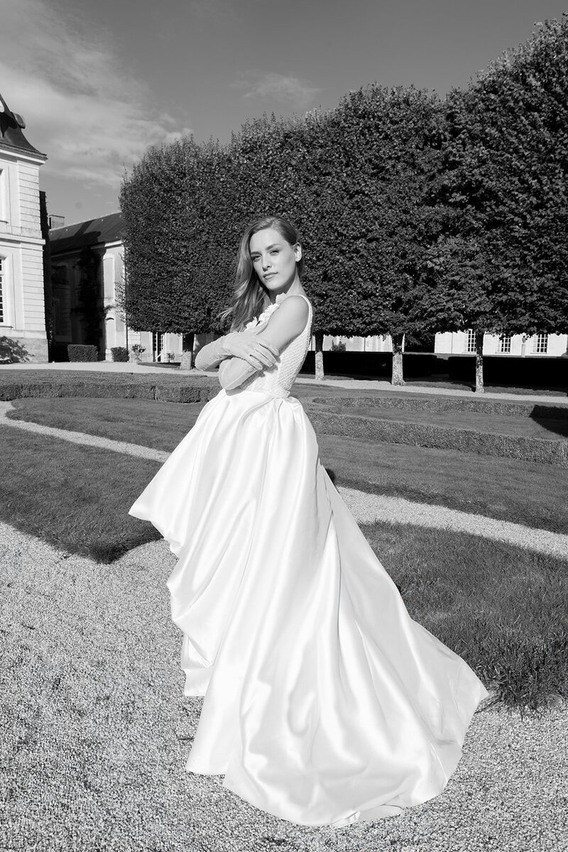 A black-and-white photo of a bride in an elegant ballgown posing in a formal garden with manicured hedges and classical architecture.