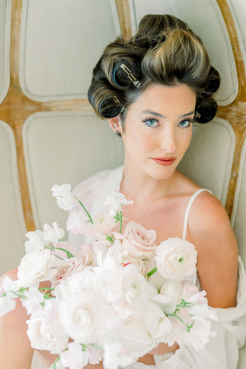 Boudoir Photographer in Richmond | A girl in white floral lace lingerie and a sheer white robe draped off her shoulders sits in a chair looking at the camera. She has curlers in her hair and has a delicate bouquet of flowers at her chest.