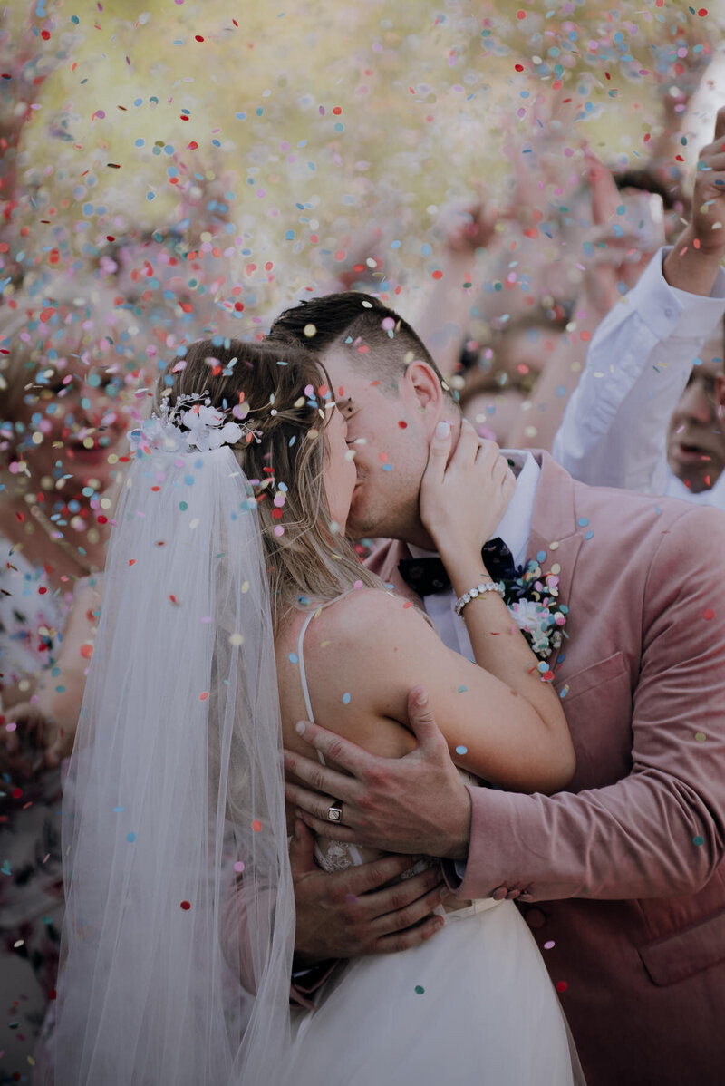 Bride and groom kissing in front of a crowd of friends with confetti flying through the air