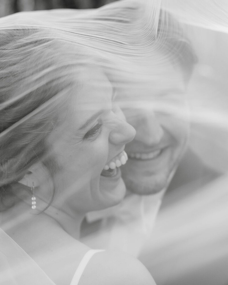 close up black and white image of smiling bride and groom on their wedding day under veil taken by wedding photographer minnesota tom thornton photography