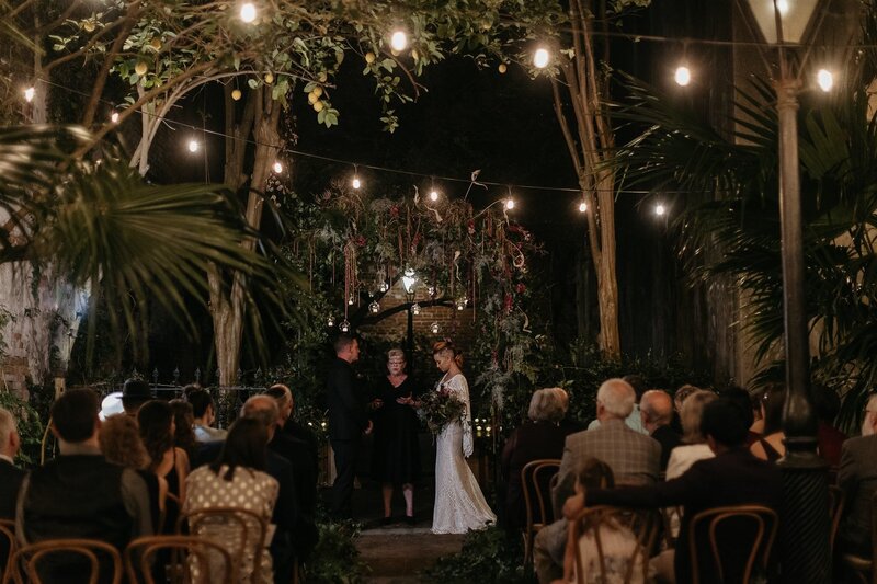 A nighttime wedding ceremony under a lush floral arch, with guests seated and watching the couple and officiant.