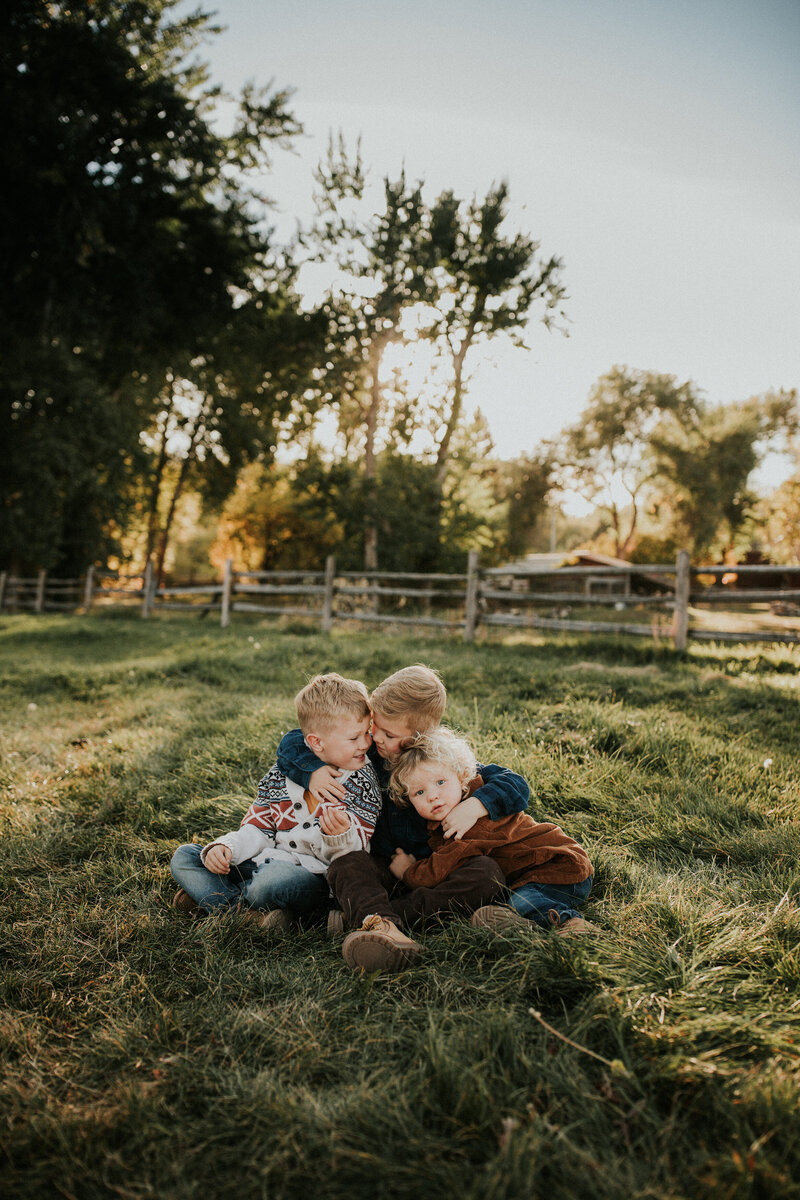 A group of three young brothers sit on the grass in a pasture with their arms wrapped around one another