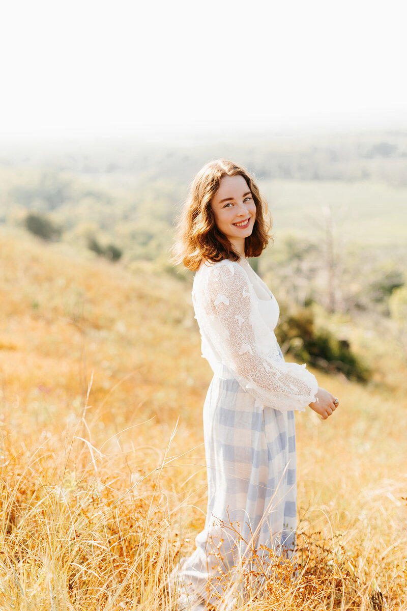 Senior girl in a white top and blue check skirt posing in a yellow grassy field