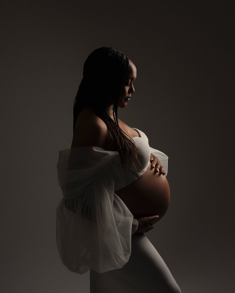 Elegant studio maternity portrait of pregnant woman in sheer off-shoulder gown with soft directional lighting in Austin, Texas.
