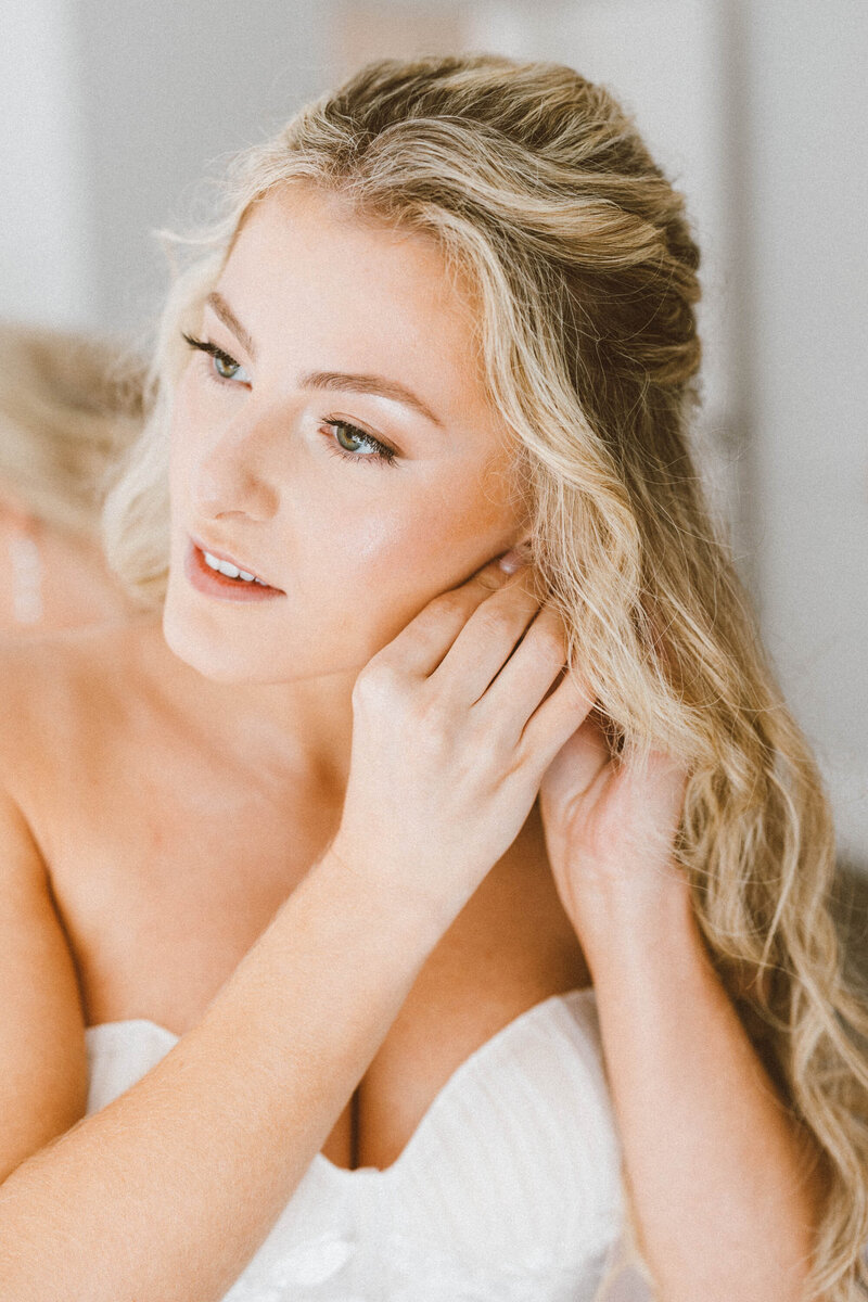 bride with blonde hair puts in earrings while gazing at herself in mirror.