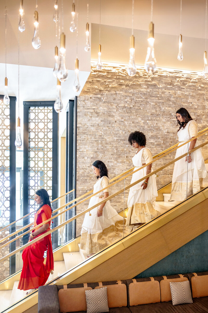 Four women in elegant dresses descend a modern staircase, with one in red leading three in white. The background features a stone wall and hanging light bulbs, creating a sophisticated atmosphere.