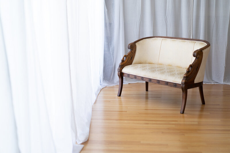 Inside the Oakland boudoir photography studio, featuring a vintage-inspired cream-colored loveseat with intricate wood carvings, set against a backdrop of sheer white curtains and warm wooden flooring.