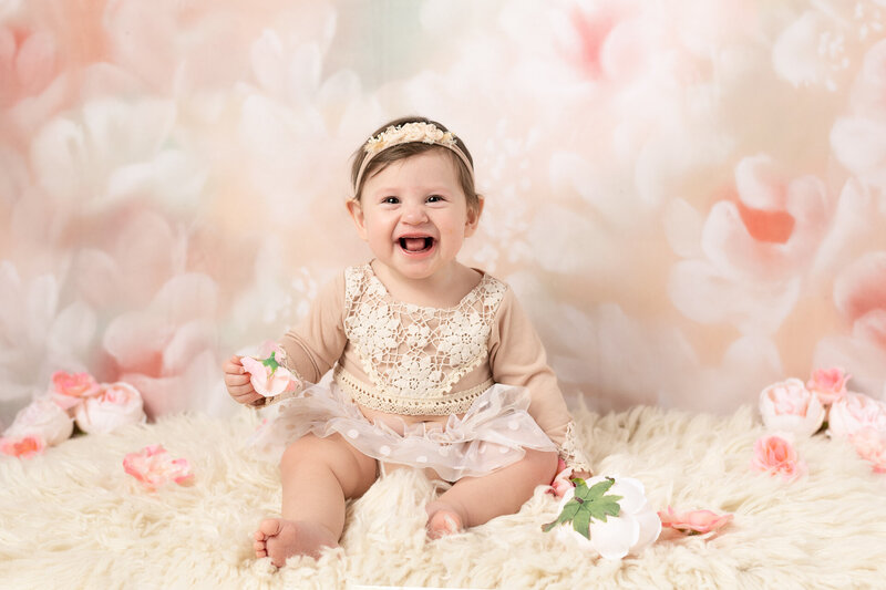 Baby milestone session — smiling baby girl in floral romper seated on white flokati with soft floral backdrop and pink flower props