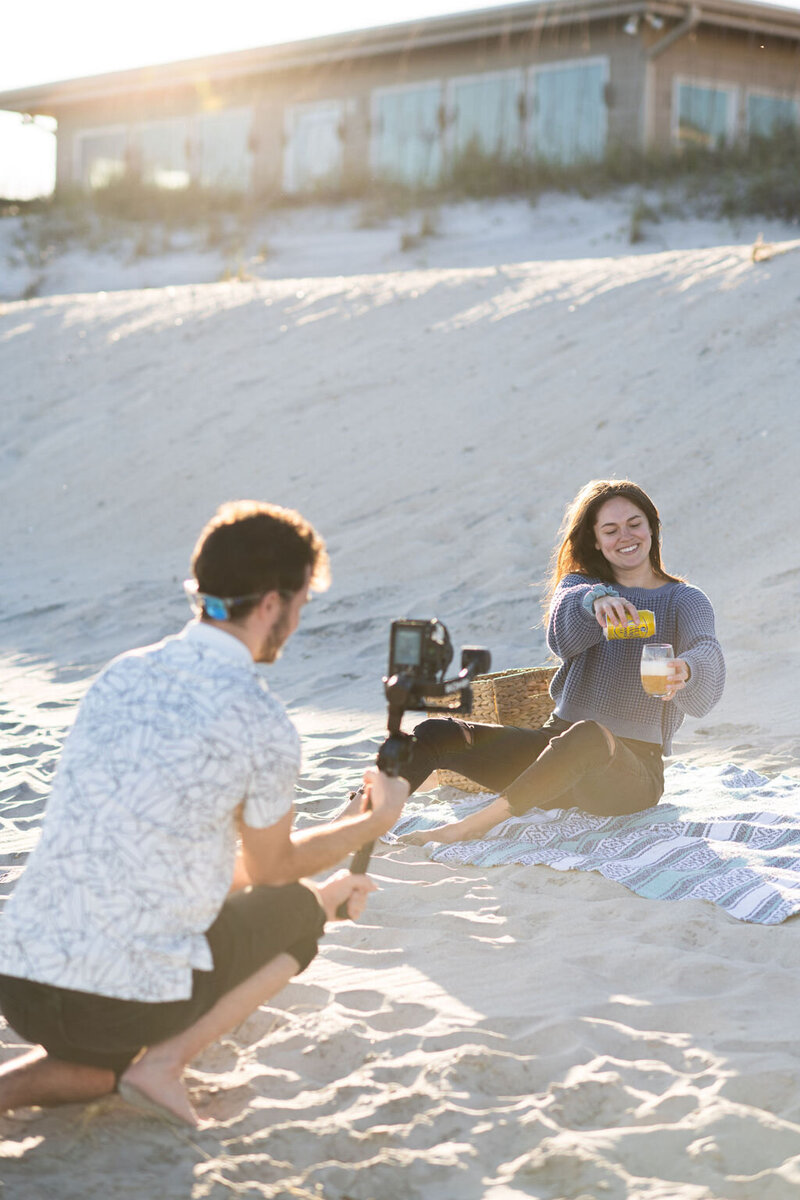 photographer holds his camera taking photos of woman sitting on the beach pouring a drink