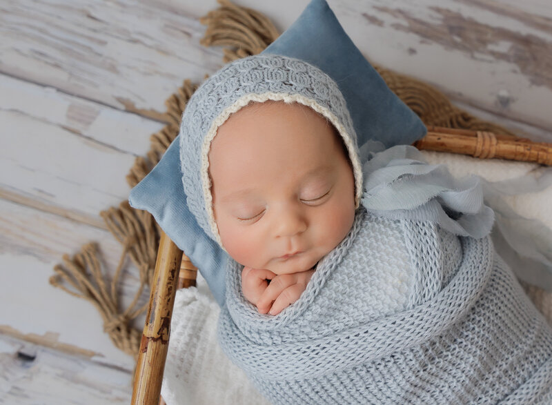 Brooklyn Newborn Photoshoot. Close up aerial image. Baby boy swaddled in a blue knit wrap and blue bonnet is sleeping peacefully  with their hands peeking out of the swaddle. Captured by best Brooklyn, NY newborn photographer Chaya Bornstein.