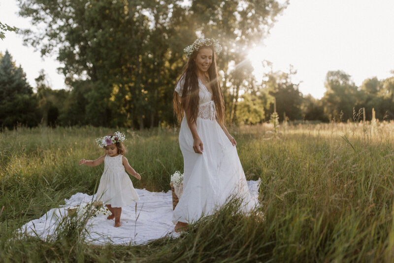 A mother and young daughter wearing white dresses and flower crowns walking barefoot in a sunlit wildflower field.