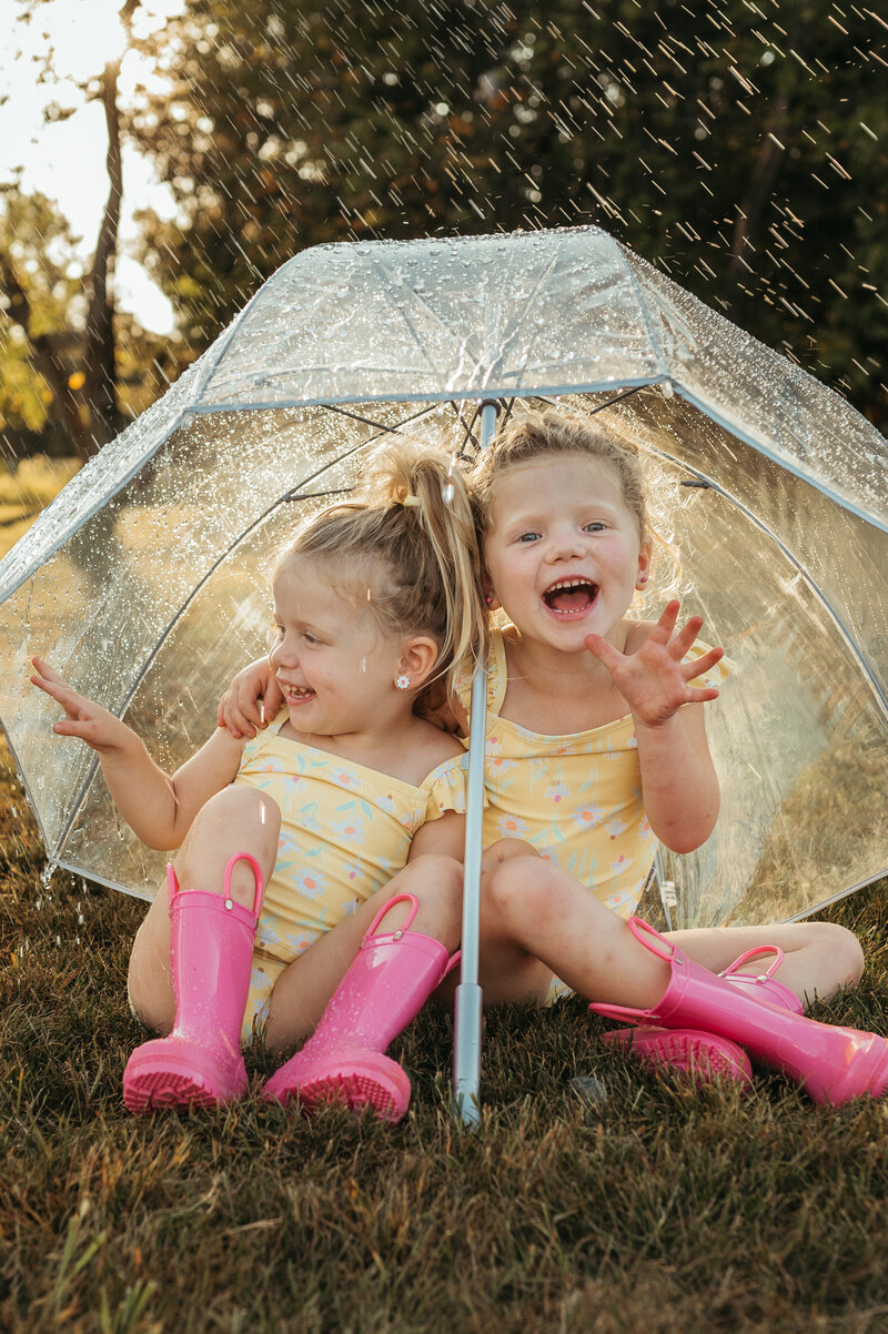 Sisters in matching pink rain boots sit under a clear umbrella  giggling in the rain. 